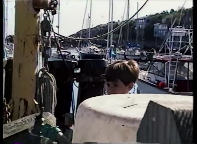 Vintage home movie footage of a young boy on a pier looking out at a scenic New England harbor full of boats and a red fishing shack