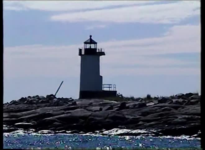 Vintage home movie footage captures a silhouetted lighthouse on a rocky coast as the ocean water sparkles