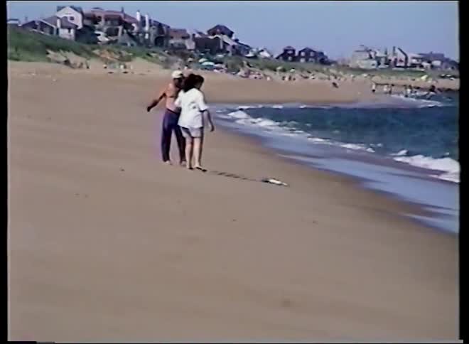 A vintage home movie captures a man and woman walking and hugging along the ocean shore on a sunny day