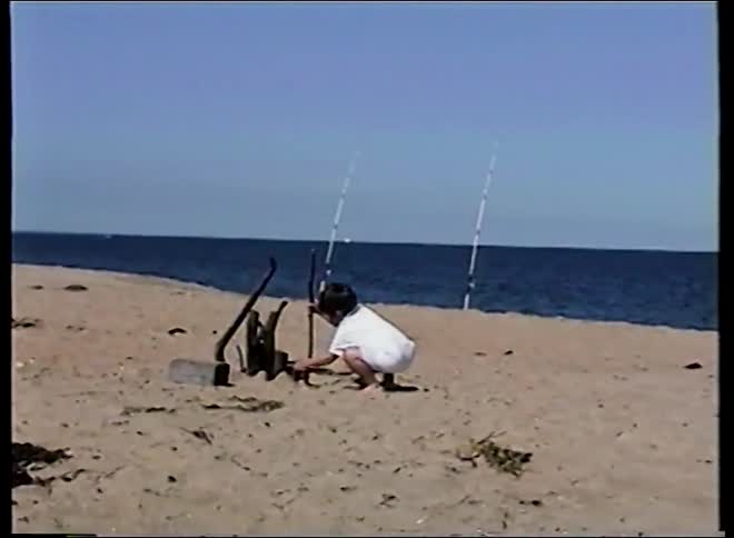 Vintage footage shows a young boy playing in the sand with fishing poles set up behind him on a sunny day at the beach