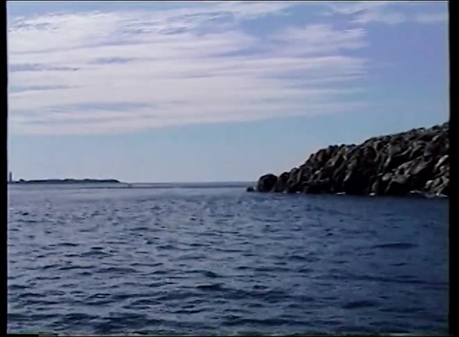 A vintage VHS shot pans from a modern house on a rocky island to a distant lighthouse across the sea