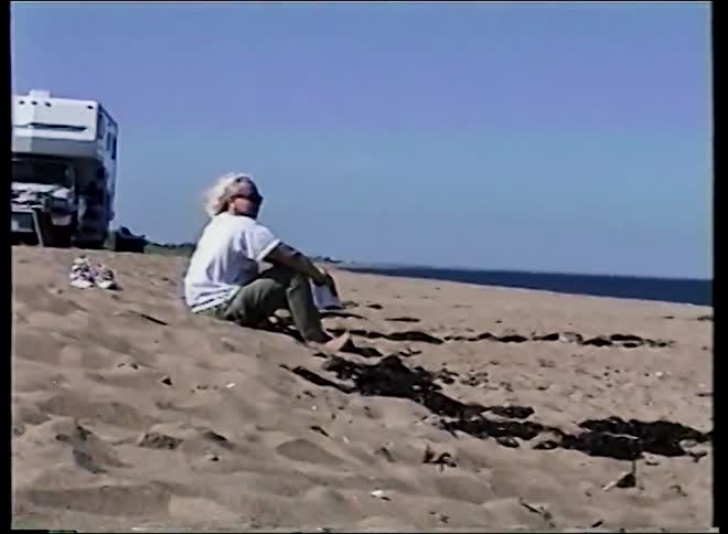 Vintage home movie captures a young boy playing near fishing poles while a woman relaxes on a sunny, sandy beach