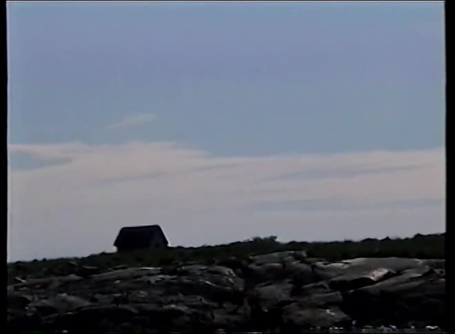 A vintage handheld shot from a boat pans from a silhouetted house to a lighthouse on a rocky shore with sparkling water in the foreground