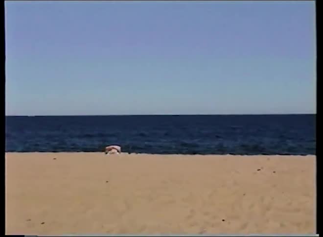 Vintage home video footage shows people relaxing on a sandy beach before the camera pans to reveal fishing rods set up by the ocean