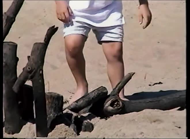 A young boy runs in slow motion on a sunny beach before stopping to play with driftwood in the sand