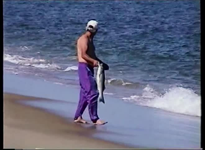 A fisherman in bright purple pants walks along the shoreline with his catch before throwing the large fish back into the ocean