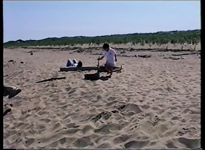 Vintage home video footage shows a young boy playing in the sand on a vast beach on a sunny day