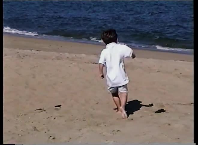 A young boy happily runs in slow motion on a sandy beach next to the ocean on a bright sunny day