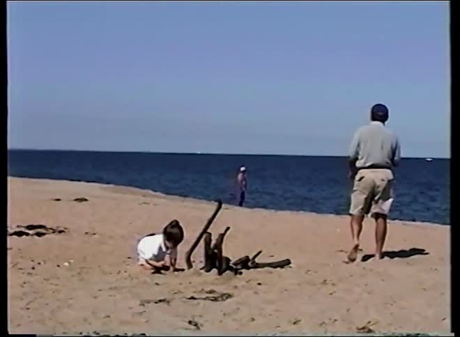 Archival footage shows a young boy playing in the sand with his grandfather on a sunny day at the beach