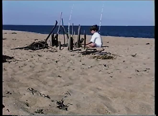 A young boy plays on a sandy beach next to fishing poles and the ocean before the camera pans to his mother sleeping on the sand next to their camper