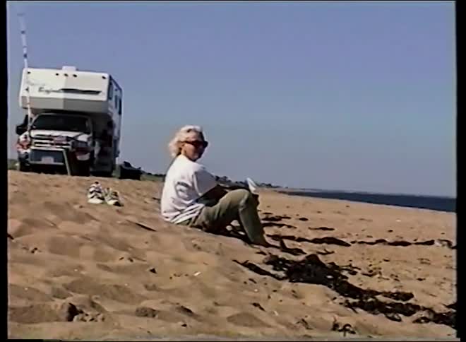 Vintage VHS footage shows a mother and her young son playing with driftwood on a sunny beach next to the ocean
