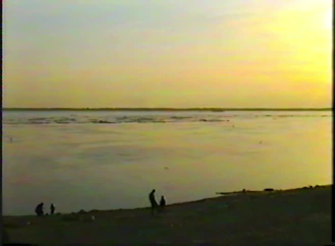 Archival footage shows silhouetted figures on the shore of a vast body of water during a tranquil sunset