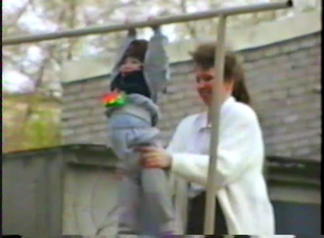 A caring mother helps her toddler hang from a horizontal bar in the backyard in this authentic vintage home video