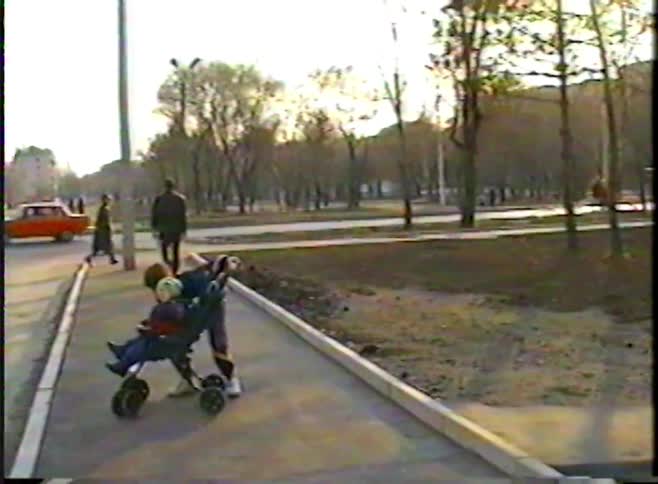 A child pushes a younger sibling in a stroller along a city sidewalk in a nostalgic vintage home video