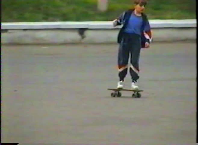 A young boy in a retro tracksuit and protective gear skillfully rides his skateboard across an empty parking lot