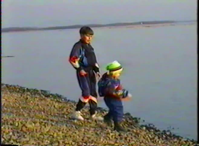 A vintage home video shows two young brothers on a rocky shore, with the older one throwing a rock into the river as the younger toddler watches