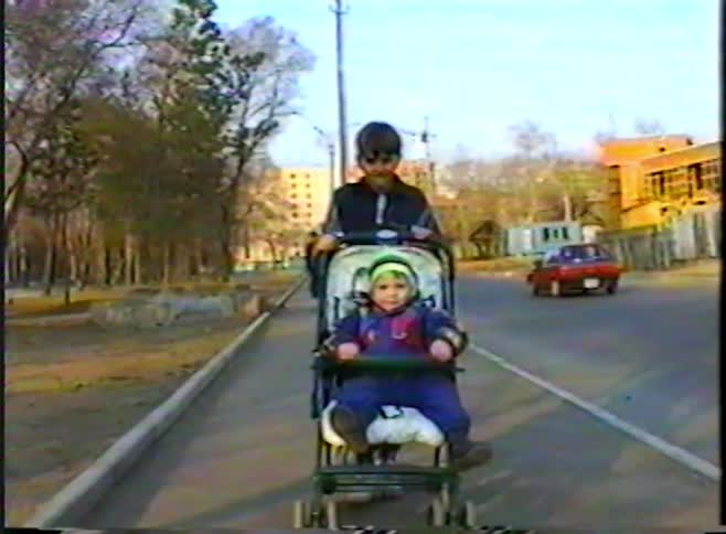 An older brother pushes his toddler sibling in a stroller along a sidewalk on a sunny day in this vintage footage
