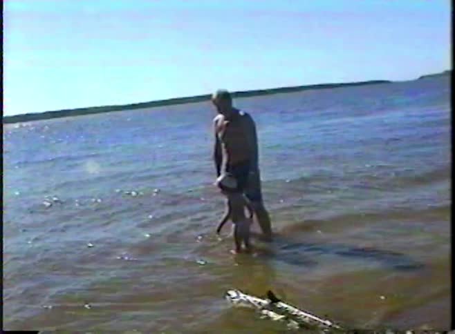 Vintage VHS footage shows a father and his young child standing in the shallow water of a lake on a sunny summer day
