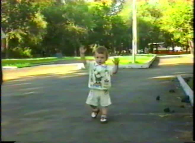 An adorable toddler joyfully claps and raises his hands while walking in a park in this nostalgic home video from the 1990s