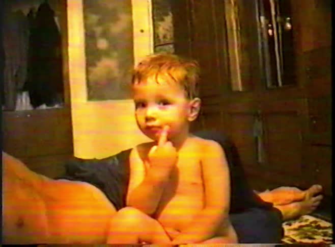 A cute, shirtless toddler looks at the camera with their finger in their mouth in a heavily distorted old home video