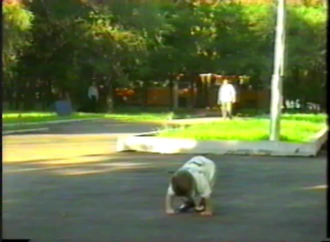 A cute toddler wobbles towards the camera before sitting down on the pavement in a sunny park in this vintage home video