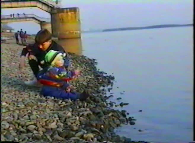 Vintage VHS footage shows two young brothers sitting together on a rocky riverbank as the toddler yawns