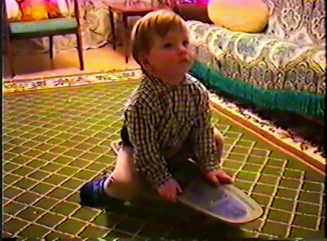Nostalgic home video of a toddler sitting on a skateboard on a patterned green rug in a vintage living room