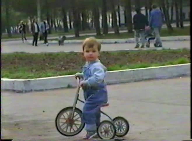 A vintage home video from the 1990s shows a small child in overalls learning to ride their first tricycle in a park