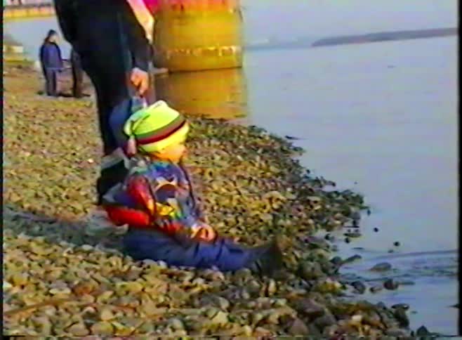 A happy toddler in a colorful winter outfit throws rocks into the river from a rocky shore in this nostalgic VHS home video