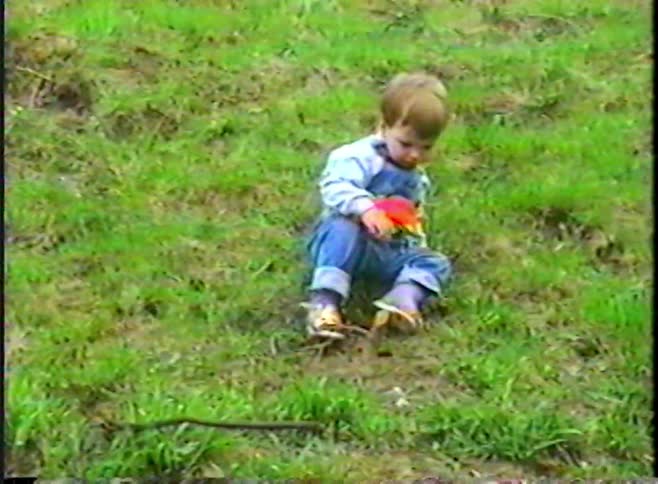 Vintage VHS footage shows a cute toddler in overalls sitting on a grassy hill, holding a red toy car and looking at the camera