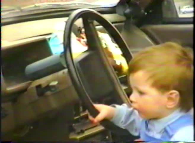 Vintage 1990s footage shows a cute toddler with red hair sitting in the driver's seat of a car and pretending to drive