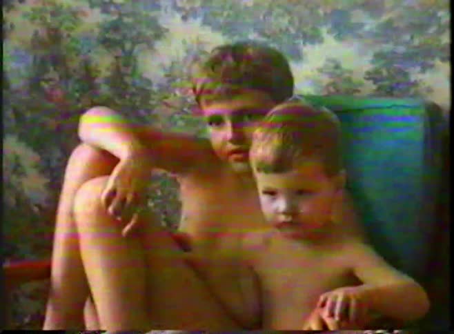 Two young children sit closely together in a chair against a patterned wall in vintage home video footage