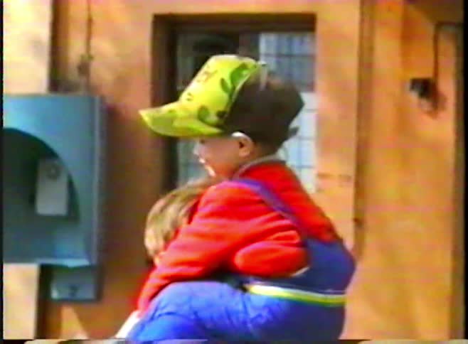 A vintage home movie captures a sweet moment of a boy carrying his younger brother on his shoulders on a city street