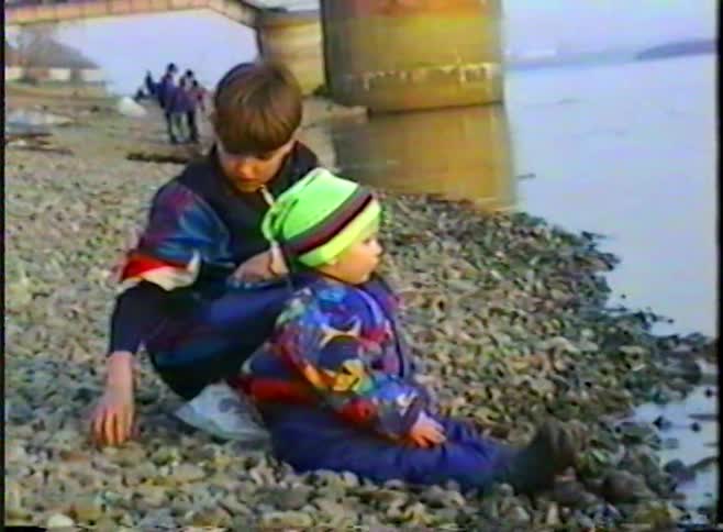 Vintage home movie footage captures two young brothers sitting on a rocky riverbank and throwing stones into the water