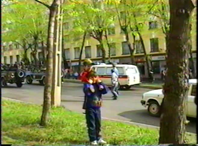 A vintage home video shows a child carrying another on their shoulders while watching a parade from the sidewalk
