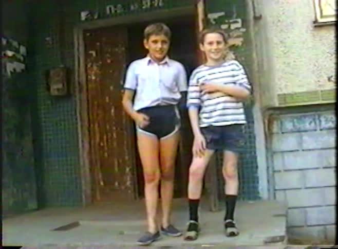 Two young boys in vintage clothing pose for a home movie camera before running inside an old building