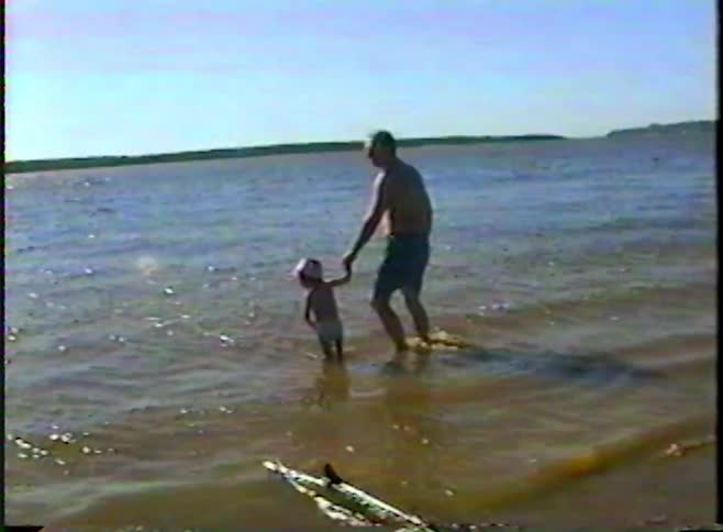 Vintage home movie captures a sweet moment of a father holding his toddler's hand as they wade in the lake on a sunny day