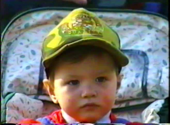 A vintage home video shows an older child pushing a toddler in a stroller through a park before zooming in for a close-up of the toddler's adorable smile
