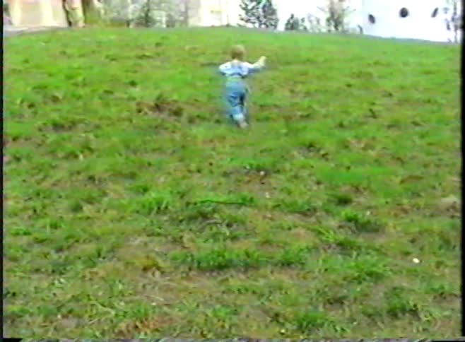Vintage 1990s footage of a cute toddler in overalls attempting to climb a grassy hill and then falling