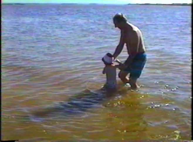 Vintage footage captures a tender moment as a grandfather helps his grandchild walk in the shallow ocean waves