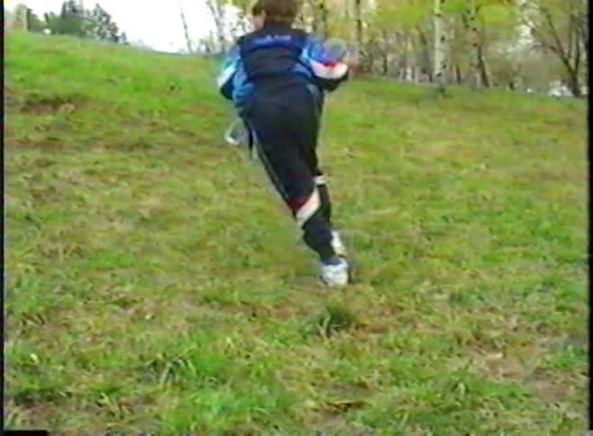 A young boy in a vintage tracksuit attempts to ride his skateboard on a grassy hill before running up with it