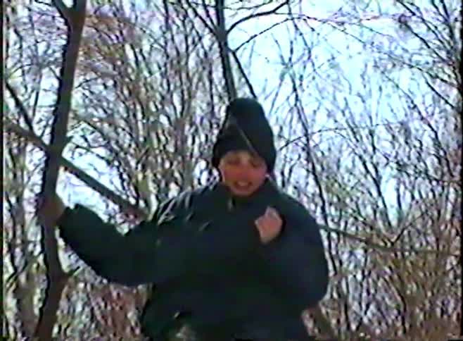 A slow zoom-in on a young boy in a winter hat and coat as he sits on a large, snow-dusted tree branch in a forest