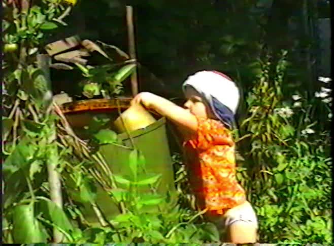 Nostalgic VHS footage shows a little girl in a sun hat playfully pouring a pot of water from a rain barrel over her head in a lush garden