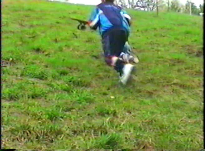 A toddler in overalls crawls and slides on a steep grassy hill while an older boy follows in this vintage home video