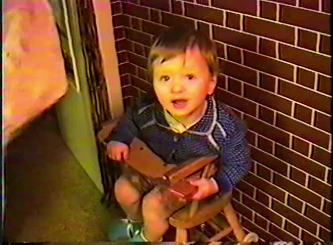A crying toddler gets a haircut from their father while sitting in a booster seat in this vintage home video