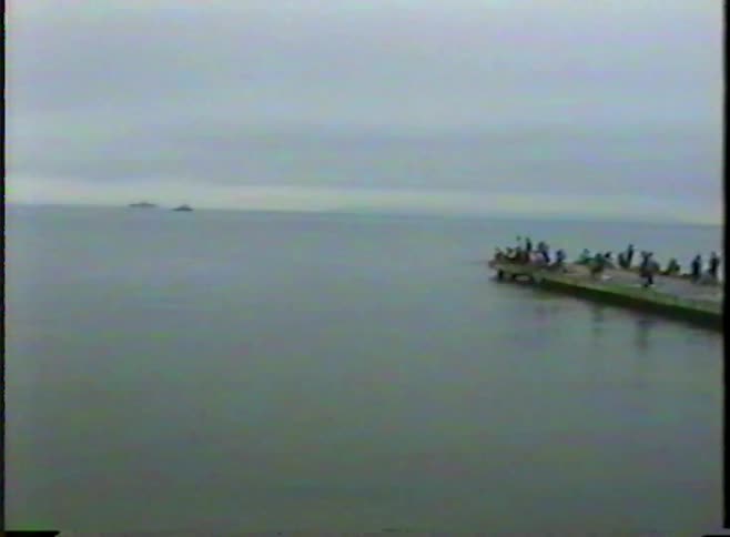 Old VHS footage pans across a vast ocean to reveal fishermen on a pier during an overcast day