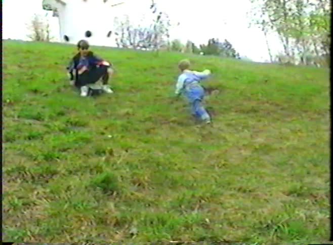 Vintage footage of a determined toddler crawling up a grassy hill as his older brother watches from his skateboard