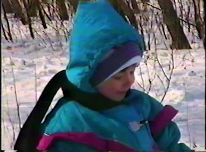 A smiling child bundled up in a colorful snowsuit sits on a log in a snowy forest in this nostalgic VHS home video clip