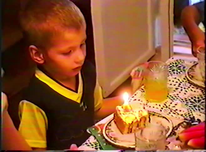 A young boy blows out a single candle on his birthday cake slice in a nostalgic home video