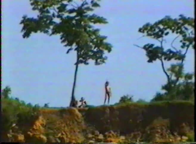 Vintage VHS footage shows a group of people standing on the edge of a high, tree-lined cliff on a sunny day